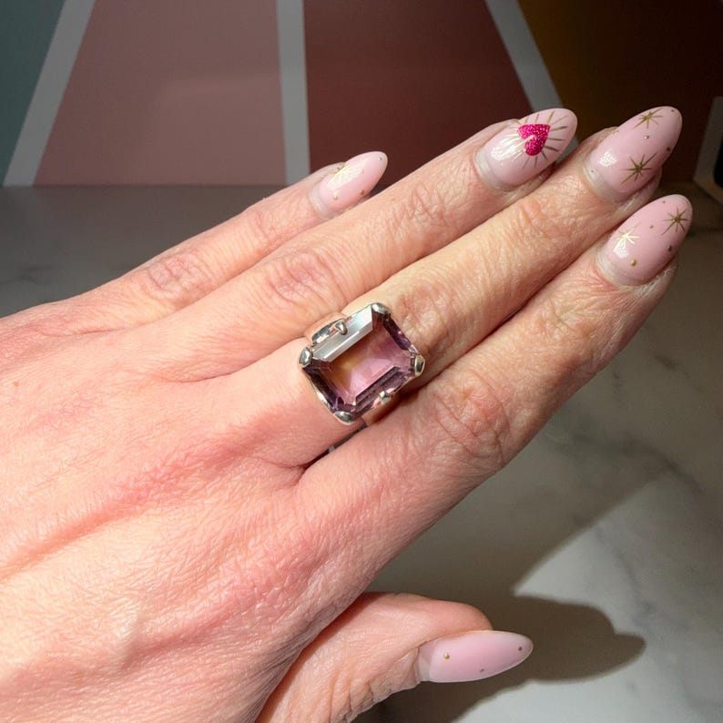 Pu&ograve; includere: Un anello d'argento con una grande ametista rettangolare, che mostra una sfumatura di colori viola e giallo. L'anello ha un'incastonatura dettagliata con elementi decorativi. L'anello &egrave; mostrato su una mano con smalto rosa.