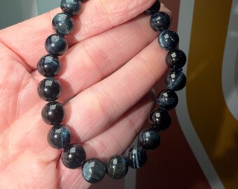 May include: A dark blue beaded bracelet. The bracelet features round, polished beads with a marbled effect. The beads are strung together on an elastic band. The bracelet is held in a hand.