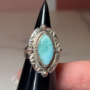 May include: A silver ring featuring an oval, pale blue gemstone. The ring has a detailed, textured design around the stone and on the band. The gemstone is a light turquoise colour. The ring is displayed on a black stand.