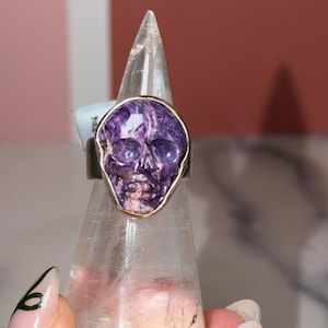 以下が含まれることがあります： 金色のアクセントが付いた、彫刻された紫色のスカルが特徴のシルバーリング。スカルリングは、透明で尖ったスタンドに展示されています。リングのデザインには、詳細な顔の特徴が含まれています。