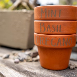 May include: Three terracotta pots stacked on top of each other. The pots are labeled "Mint", "Basil", and "Oregano".