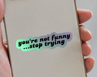 以下が含まれることがあります： 「you're not funny...stop trying」という黒い文字が書かれたホログラフィック仕上げのステッカーです。