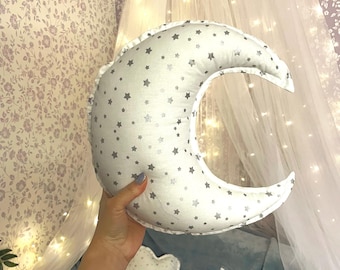 May include: A white crescent moon-shaped pillow with silver stars printed on it. The pillow is being held by a hand in front of a white sheer canopy with string lights.