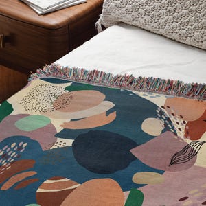May include: A colorful throw blanket with a geometric pattern of abstract shapes in shades of blue, green, pink, and orange. The blanket has a fringed edge.