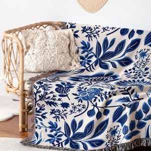 Puede incluir: Una manta tejida con un estampado floral azul está sobre un sofá de ratán. También se ven un cojín de macramé y un tapiz tejido. La manta tiene un fondo crema con hojas y flores azules. El sofá tiene un cojín crema.