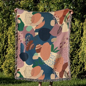 May include: A woven throw blanket with an abstract pattern featuring various shapes and colors, including blue, green, pink, orange, and beige. The blanket has a fringed edge.