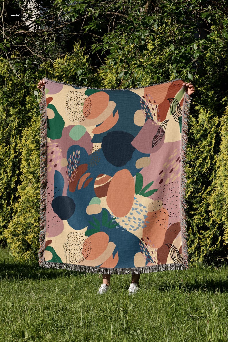 May include: A woven throw blanket with an abstract pattern featuring various shapes and colors, including blue, green, pink, orange, and beige. The blanket has a fringed edge.