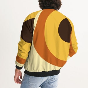 May include: A black bomber jacket with a retro-style geometric pattern in shades of yellow, orange, and brown. The pattern features concentric circles and arcs.