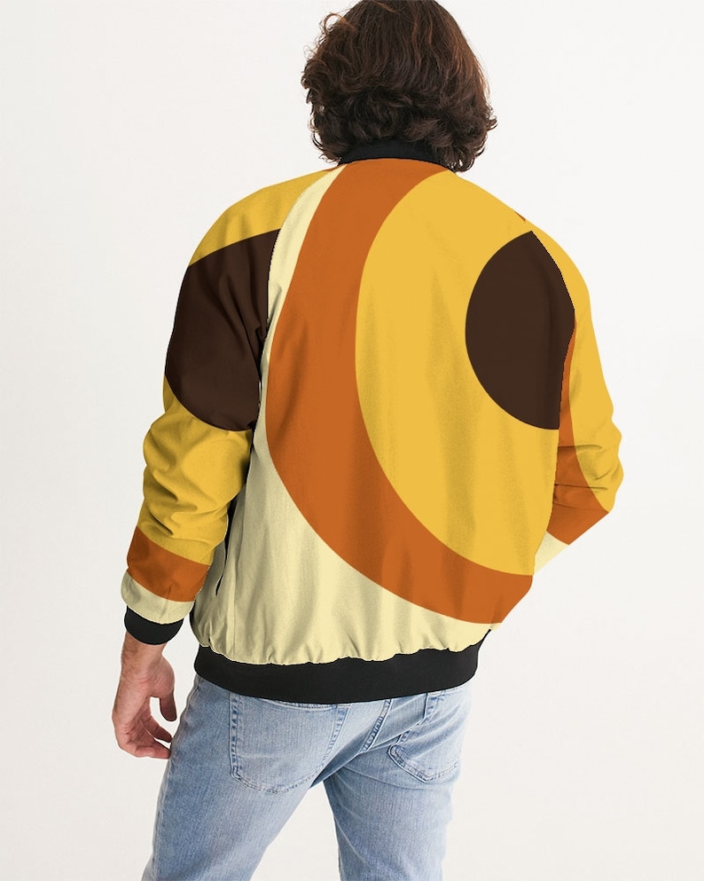 May include: A black bomber jacket with a retro-style geometric pattern in shades of yellow, orange, and brown. The pattern features concentric circles and arcs.