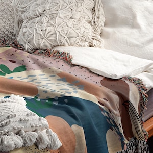 May include: A colorful abstract throw blanket with fringe edges, featuring a mix of blue, brown, beige, and green colors. The blanket is draped over a couch with white pillows.