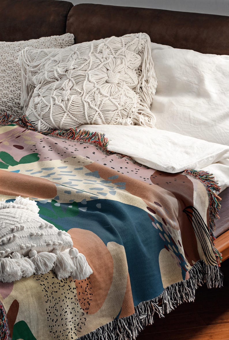 May include: A colorful abstract throw blanket with fringe edges, featuring a mix of blue, brown, beige, and green colors. The blanket is draped over a couch with white pillows.