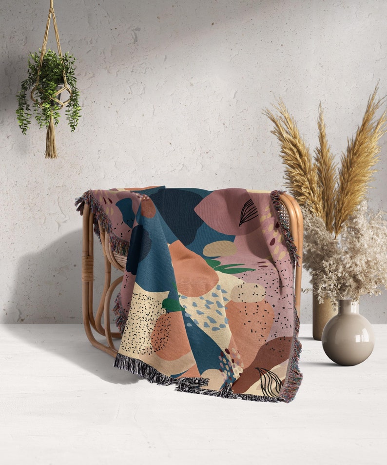 A cotton blanket with an abstract design in mauve, dark teal blue, peach and chestnut tones, thrown over a cane chair. There are vases with dried flowers sitting net to the chair and a hanging plant to the left of the picture