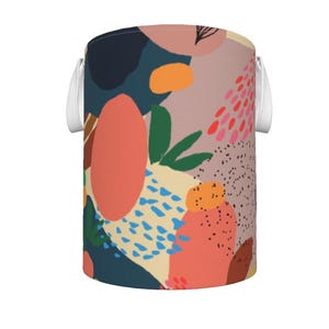Puede incluir: Un cubo de almacenamiento cilíndrico con un colorido patrón abstracto. El diseño presenta formas grandes en coral, azul marino y beige, con acentos de verde, azul y negro. Las asas blancas están unidas a los lados.