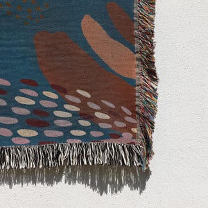 May include: A woven throw blanket with a blue background and abstract shapes in shades of brown, pink, and white. The blanket has a black and white fringe.