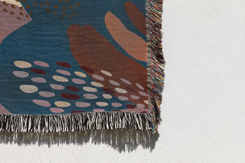 May include: A woven throw blanket with a blue background and abstract shapes in shades of brown, pink, and white. The blanket has a black and white fringe.