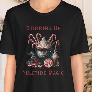 May include: Black t-shirt with a red graphic of a cauldron with candy canes, whipped cream, and a candy cane lollipop. The text "Stirring Up Yuletide Magic" is printed in red below the graphic.