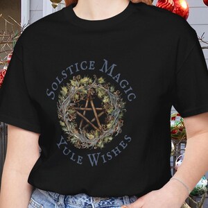 May include: Black t-shirt featuring a pentagram design within a wreath of leaves and berries. The text "SOLSTICE MAGIC YULE WISHES" is arched around the wreath. The shirt is made of a soft material and is suitable for casual wear.
