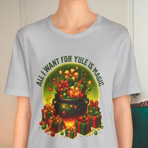 May include: A light gray t-shirt featuring a colorful graphic. The graphic shows a cauldron overflowing with wrapped gifts and a banner that reads "ALL I WANT FOR YULE IS MAGIC". The design includes green, red, and gold elements.