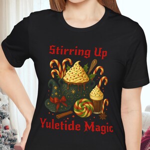 May include: Black t-shirt with a festive graphic. The design features a witch's hat, candy canes, and a swirl of whipped cream, with the text "Stirring Up Yuletide Magic" in red. The shirt is a crew neck.