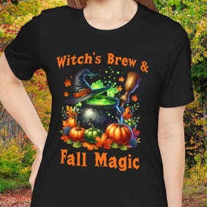 May include: Black t-shirt with the text "Witch's Brew & Fall Magic" in orange. The graphic features a witch's hat, cauldron with green liquid, pumpkins, and a broom. The design is perfect for Halloween.