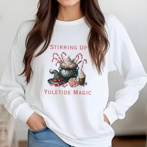 May include: White long-sleeve shirt with a graphic featuring a cauldron, candy canes, and a witch's hat. The text "Stirring Up Yuletide Magic" is printed above and below the graphic. The design includes a swirl of whipped cream and a chocolate drink.