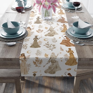 May include: White table runner with a repeating pattern of watercolor figures in flowing gowns, crescent moons, and botanical elements in shades of brown and gold. The runner is placed on a wooden table set for a meal, with plates, bowls, and wine glasses.