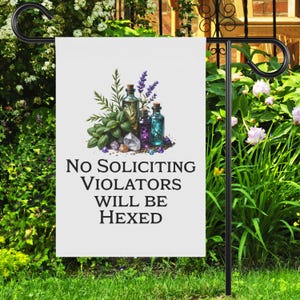 May include: A white garden flag with the text "No Soliciting Violators Will Be Hexed." The flag features an illustration of potion bottles, crystals, and herbs. The flag is displayed on a black metal garden stake, surrounded by green plants and flowers.