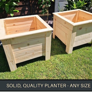 May include: Two wooden planter boxes made of natural wood. The boxes are square and have a simple design. The boxes are sitting on a grassy surface.