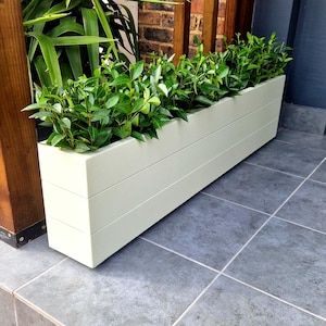 May include: A long, cream-coloured rectangular planter box filled with green plants. The box has a slatted design and is on a tiled surface. The planter is next to a wooden post and a brick wall.