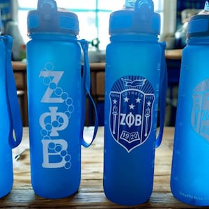 May include: Four translucent blue plastic water bottles with dark blue lids and carrying straps. Each bottle features white Greek lettering ZΦB and the year 1920, with varying graphic designs including a pearl strand and a crest emblem.