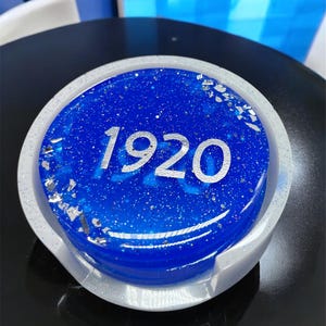 May include: A white coaster with a blue and silver glitter circle in the center. The circle has the number "1920" printed in silver.