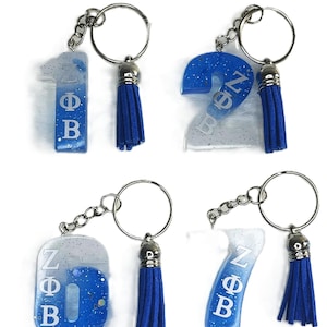 May include: Four keychains with silver-tone hardware, blue tassels, and resin numbers 1, 2, 6, and 7. Each number is in a blue and white glitter design with the Greek letters ZΦB.