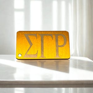 May include: A rectangular gold-toned acrylic tag featuring the Greek letters Sigma Gamma Rho in a cutout style. The tag has a smooth, reflective surface and includes a small circular hole on the bottom left corner for attachment purposes.