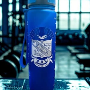 May include: A translucent blue water bottle with a gray lid and a blue carrying strap. The front features a white graphic emblem with a bird, a shield, and the Greek letters ΦΒΣ above the text FOUNDED 1914. Measurement markings are visible on the side.