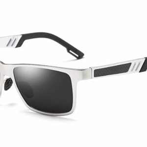 Puede incluir: Gafas de sol plateadas con lentes oscuras. Las gafas tienen forma rectangular con detalles negros en las patillas y las varillas. Las lentes son oscuras y la montura es de color plateado brillante.