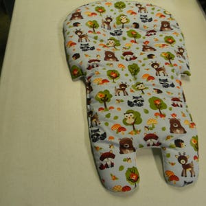 Può includere: Un inserto imbottito per seggiolino per bambini con sfondo azzurro. L'inserto è ricoperto da un motivo ripetuto di animali del bosco, alberi e funghi in vari colori. L'inserto ha una forma unica con ritagli.
