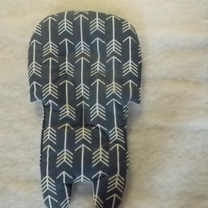 May include: A gray and white patterned baby pillow with a repeating arrow design. The pillow is rectangular with a rounded top and two small, rounded extensions at the bottom. The pillow is on a white, textured surface.