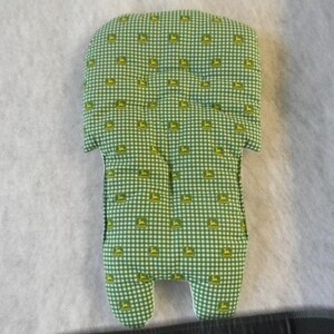 Può includere: Inserto per seggiolino auto per bambini con motivo a quadretti verde e bianco con logo giallo ripetuto. L'inserto ha una parte superiore arrotondata, due alette laterali e due prolungamenti a forma di piede nella parte inferiore. Il tessuto è un materiale morbido e trapuntato.