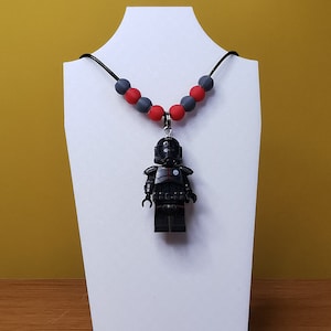 May include: A black Lego minifigure of a Star Wars character wearing a black helmet and armor, hanging from a necklace with red and gray beads.