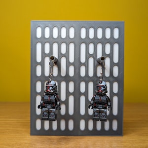 May include: A pair of silver earrings featuring a black and red Lego minifigure of a Star Wars clone trooper. The earrings are hanging from a gray display stand with a grid pattern.