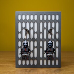 May include: A pair of black and red Star Wars themed earrings featuring a Lego minifigure of a Stormtrooper. The earrings are hanging from a grey display stand with a white grid pattern.