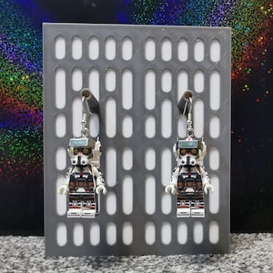 Può includere: Un paio di orecchini con minifigure Lego bianche e nere con un design di clone trooper di Star Wars. Gli orecchini sono esposti su uno sfondo a griglia grigia.