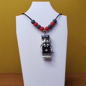 Könnte beinhalten: Eine schwarze Halskette mit roten und grauen Perlen und einer Lego-Minifigur eines Star Wars Klon-Soldaten, die daran hängt.
