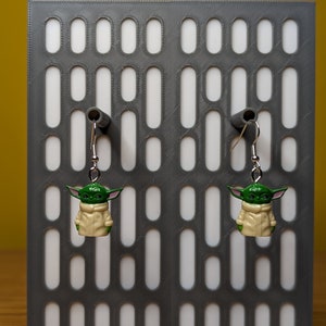 May include: A pair of silver dangle earrings with green and beige character charms. The charms are of a fictional character with large ears and a hooded robe.