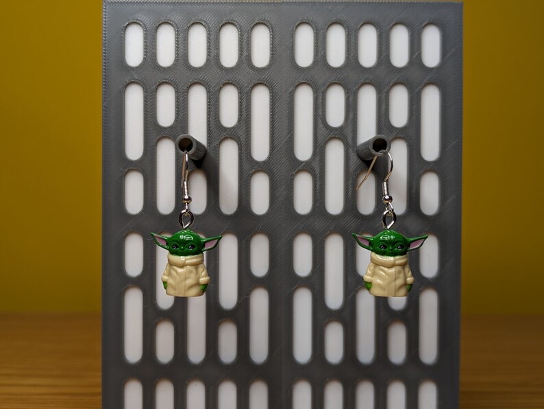 May include: A pair of silver dangle earrings with green and beige character charms. The charms are of a fictional character with large ears and a hooded robe.