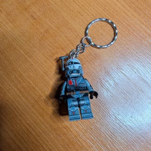 Pode incluir: Um porta-chaves com uma pequena figura de um personagem em uniforme cinza e azul, completo com capacete e um porta-chaves prateado. A figura tem uma ferramenta presa ao capacete. O porta-chaves está sobre uma superfície de madeira.