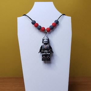 Könnte beinhalten: Eine schwarze und rote Perlenkette mit einem Lego-Anhänger von Darth Vader.
