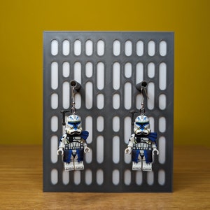 May include: A pair of earrings featuring miniature Lego figures of a clone trooper, with white, blue, and black details. The earrings hang from silver hooks against a gray display board with a pattern of oval cutouts.