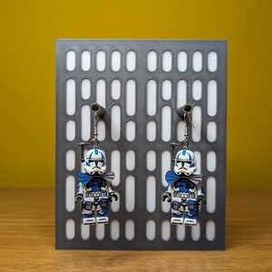 May include: A pair of blue and white Lego Star Wars clone trooper earrings hanging on a gray display stand. The earrings feature a blue and white clone trooper with a blue bow tie.