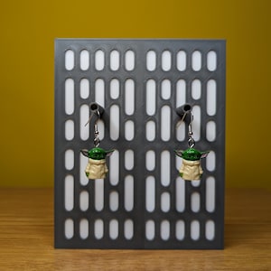 May include: A pair of dangle earrings featuring a green and beige character with large ears. The earrings are hanging from a gray display stand with a white background.